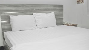 Una cama bien hecha con sábanas y almohadas blancas, un cabecero de madera y una mesita de noche con un teléfono y un objeto pequeño encima.