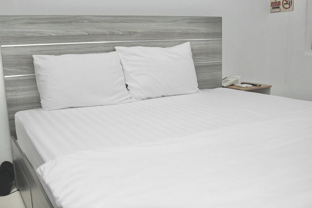 A neatly made bed with white linens and pillows, a wooden headboard, and a bedside table with a phone and a small item on it.