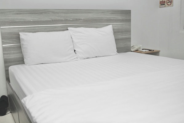 Una cama bien hecha con sábanas y almohadas blancas, un cabecero de madera y una mesita de noche con un teléfono y un objeto pequeño encima.