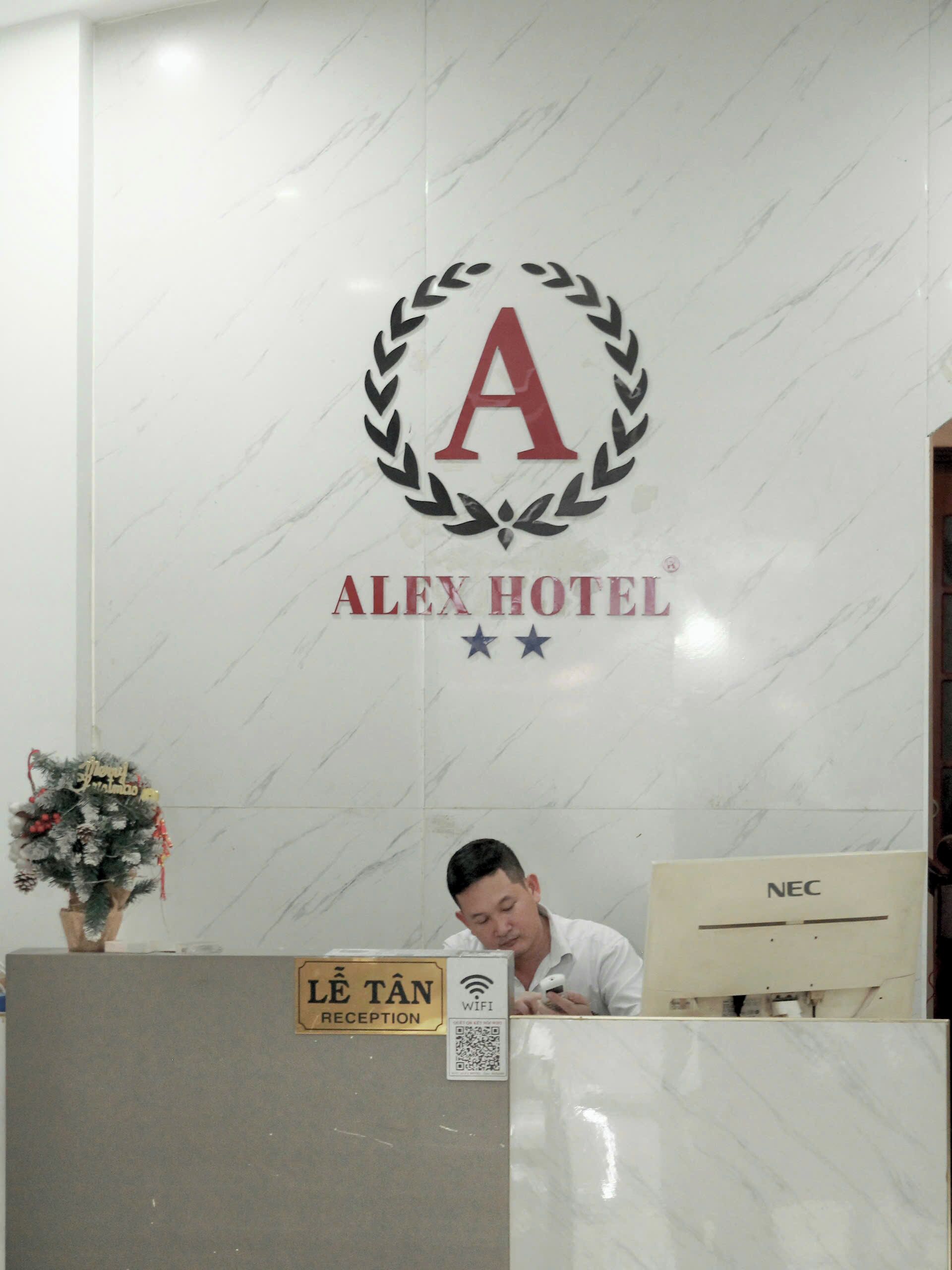 A reception desk with a marble wall, a logo for 'ALEX HOTEL,' and a sign indicating 'LE TÂN RECEPTION.'