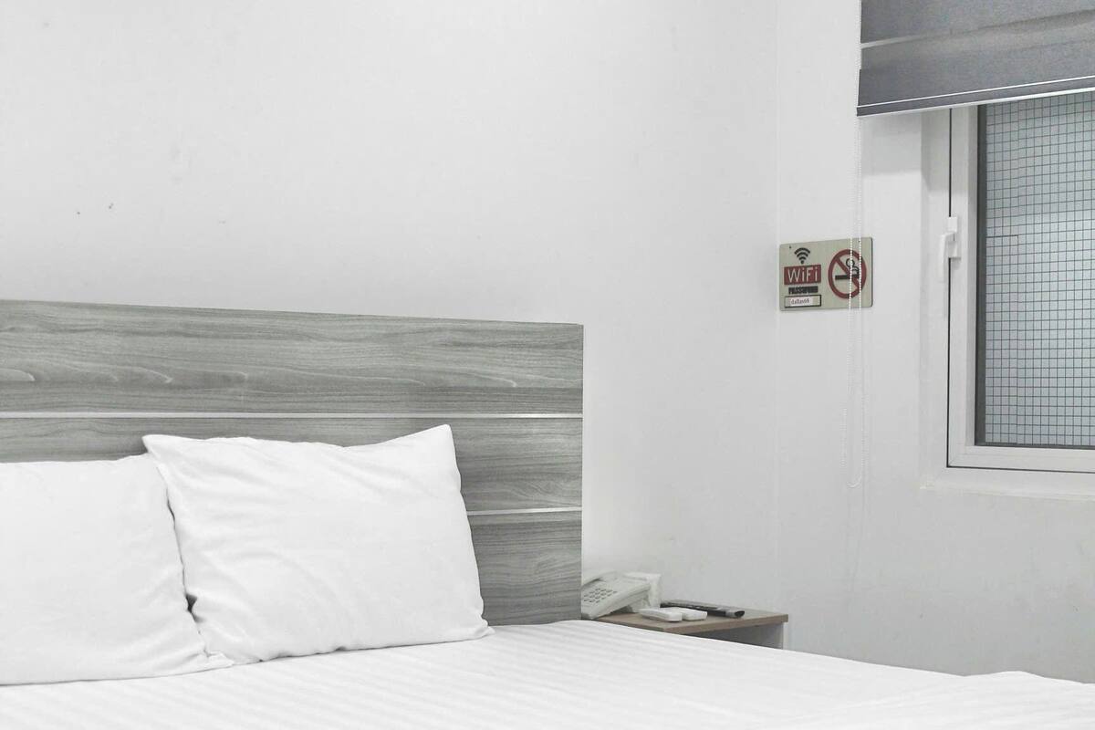A hotel room with a bed, a bedside table with a phone, and a window with a grid pattern.