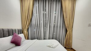 A neatly made bed with white linens and a single pink pillow, a grey headboard, and sheer curtains with a city view. - Vinhome Skylake Luxury Apartment  (Hanoi)
