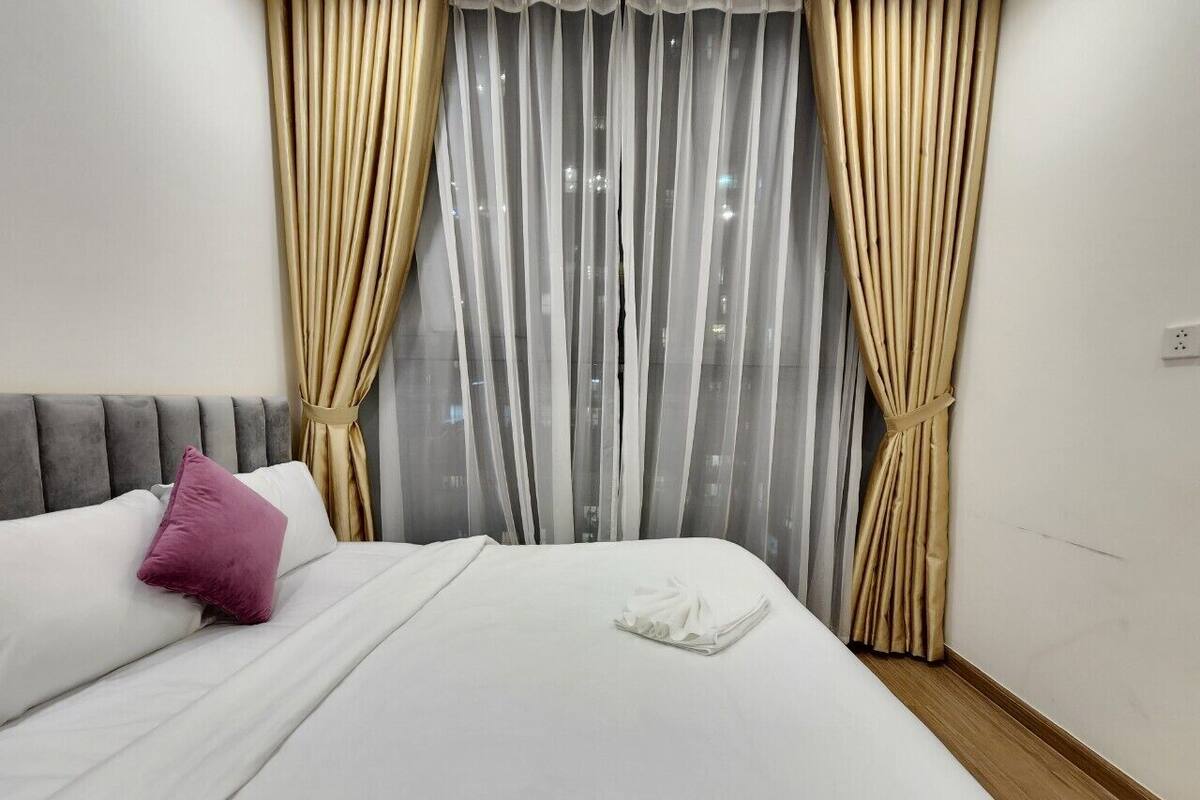 Cama bem arrumada com lençóis brancos e um único travesseiro rosa, cabeceira cinza e cortinas leves com vista para a cidade.
