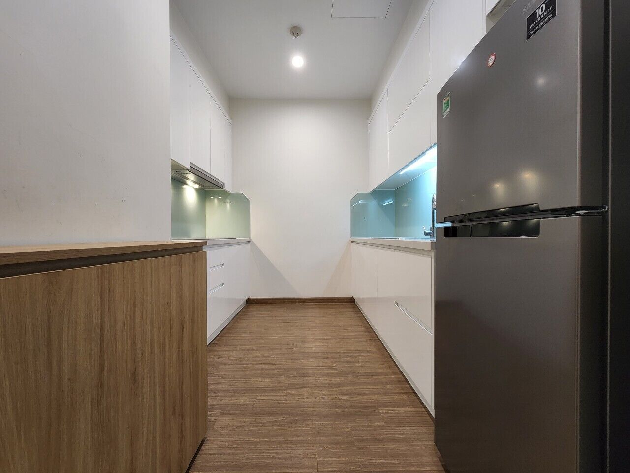 Un couloir étroit doté d’un plancher en bois, de murs blancs et d’un réfrigérateur en acier inoxydable, situé sur le côté droit.
