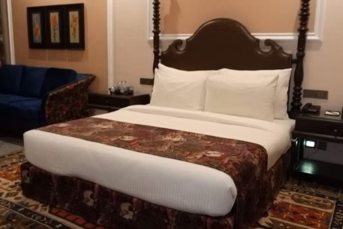 A neatly made bed with a patterned bedspread, a dark wooden headboard, and white pillows. There is a blue armchair and a small table with a lamp in the room.