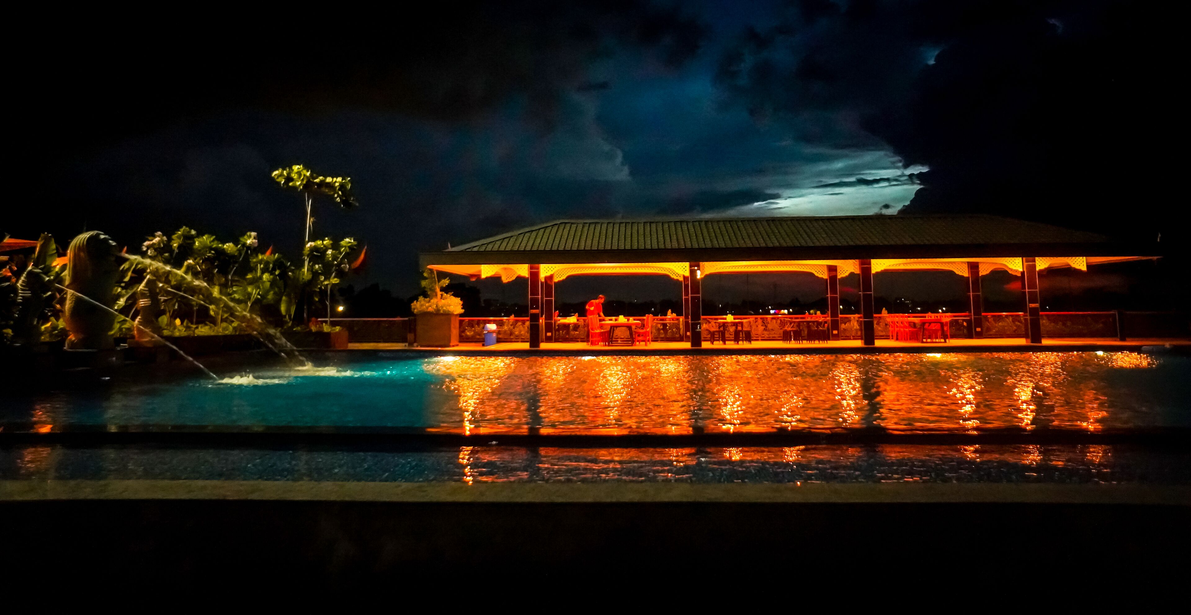 Zona de piscina nocturna con área de estar cubierta y una gran fuente de agua.