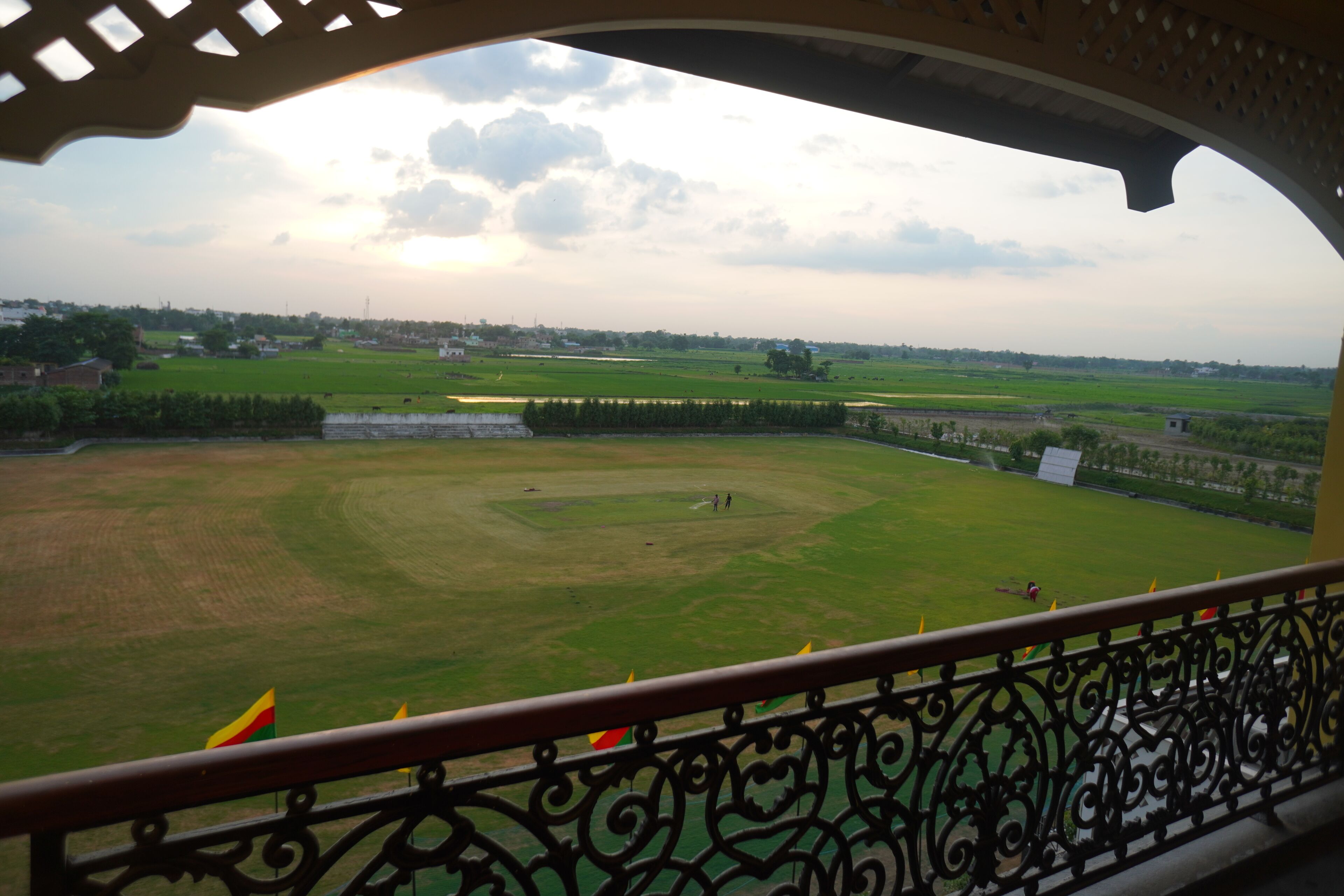 A view from a balcony with a wrought iron railing, overlooking a large green field with a few flags, and a distant town.