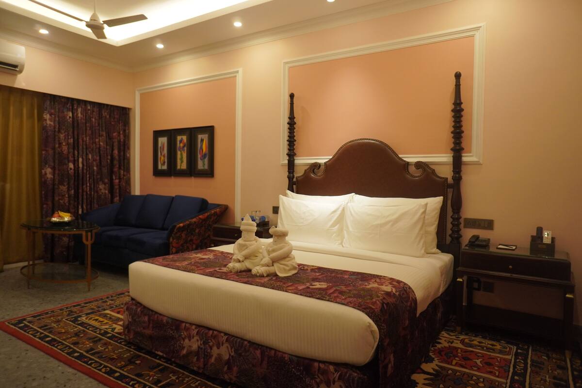 A hotel room with a four-poster bed, a blue armchair, a small table with a lamp, and a decorative rug.