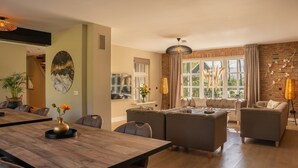 A spacious living room with a large sofa set, a dining area with a wooden table, and a view of the outdoors through large windows. - Group House in Twente with Hot Tub (Losser)