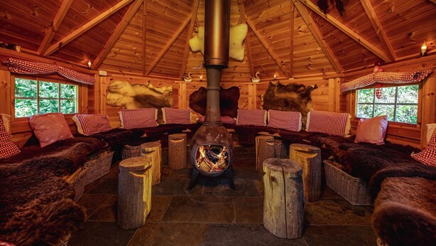 A cozy cabin interior with a central wood-burning stove, wooden walls, and checkered cushions. - Group House in Twente with Hot Tub (Losser)