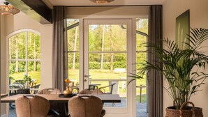 A dining area with a table and chairs, a large window with curtains, and a potted plant. - Group House in Twente with Hot Tub (Losser)