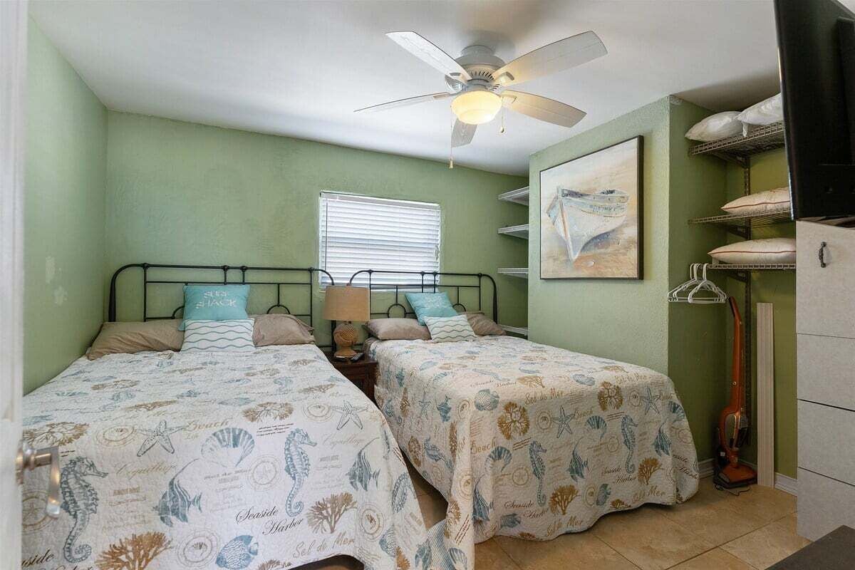 A bedroom with two beds, a ceiling fan, a window with blinds, a painting, and a vacuum cleaner.