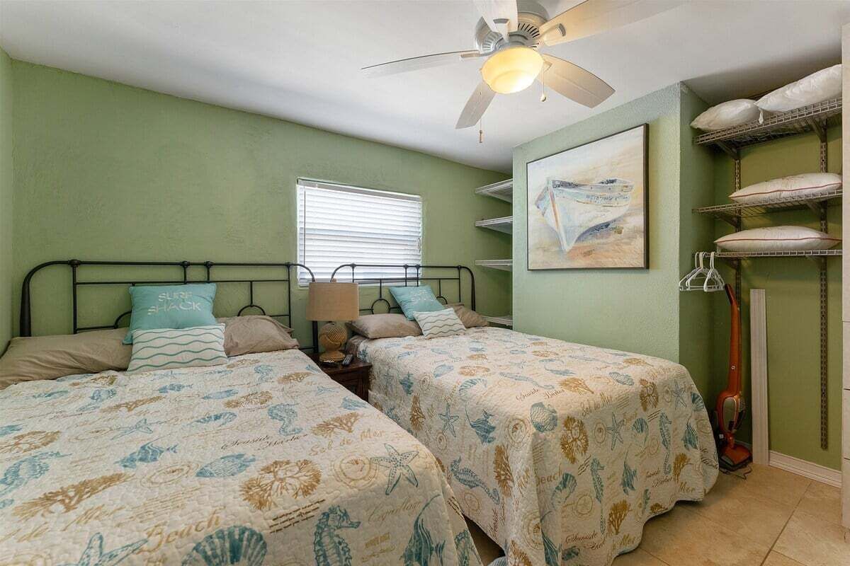 Une chambre avec deux lits, un ventilateur de plafond, une fenêtre avec des stores, un tableau au mur et une étagère avec des coussins.