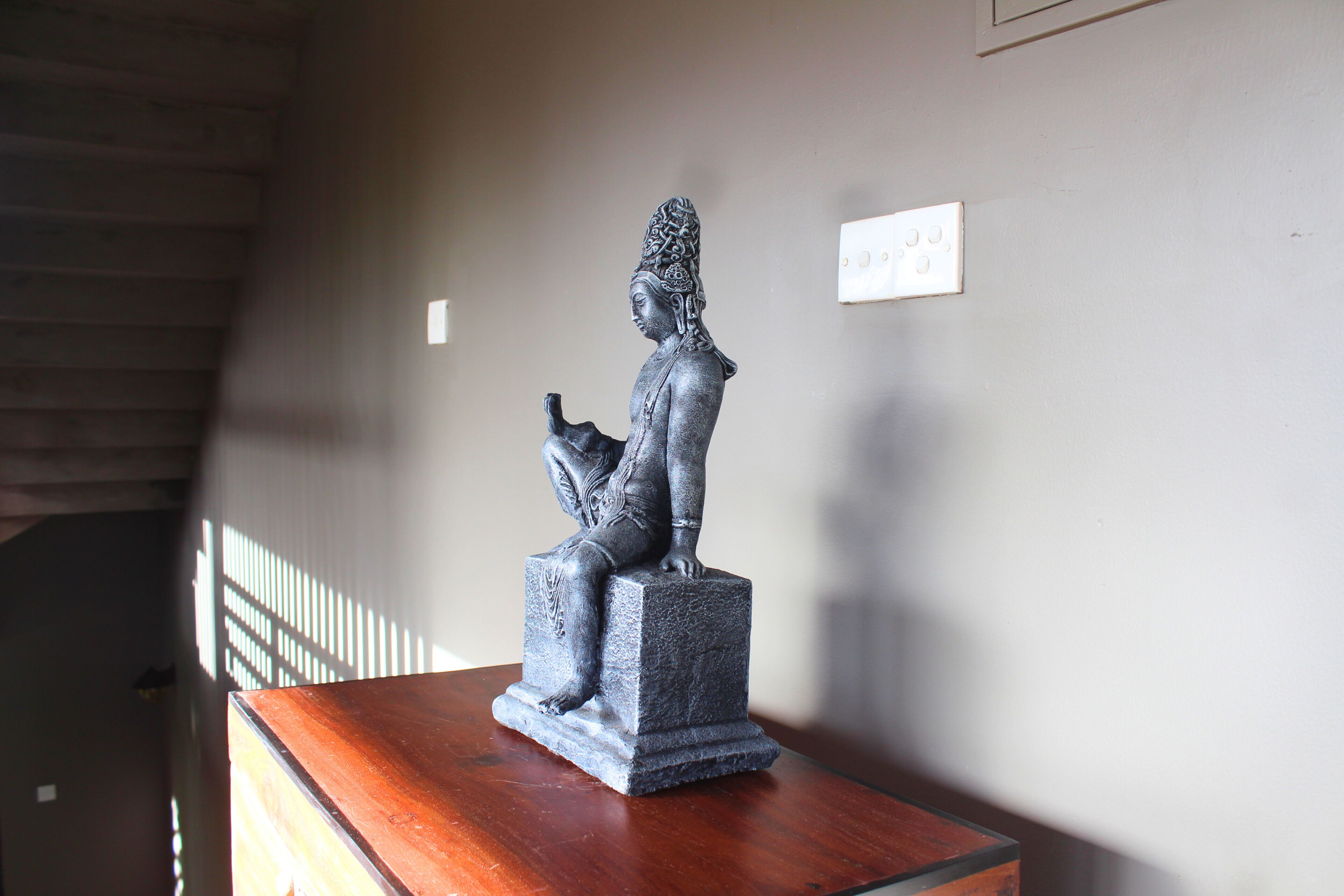 A statue of a seated figure on a pedestal in a room with a sloped ceiling and a light switch on the wall.