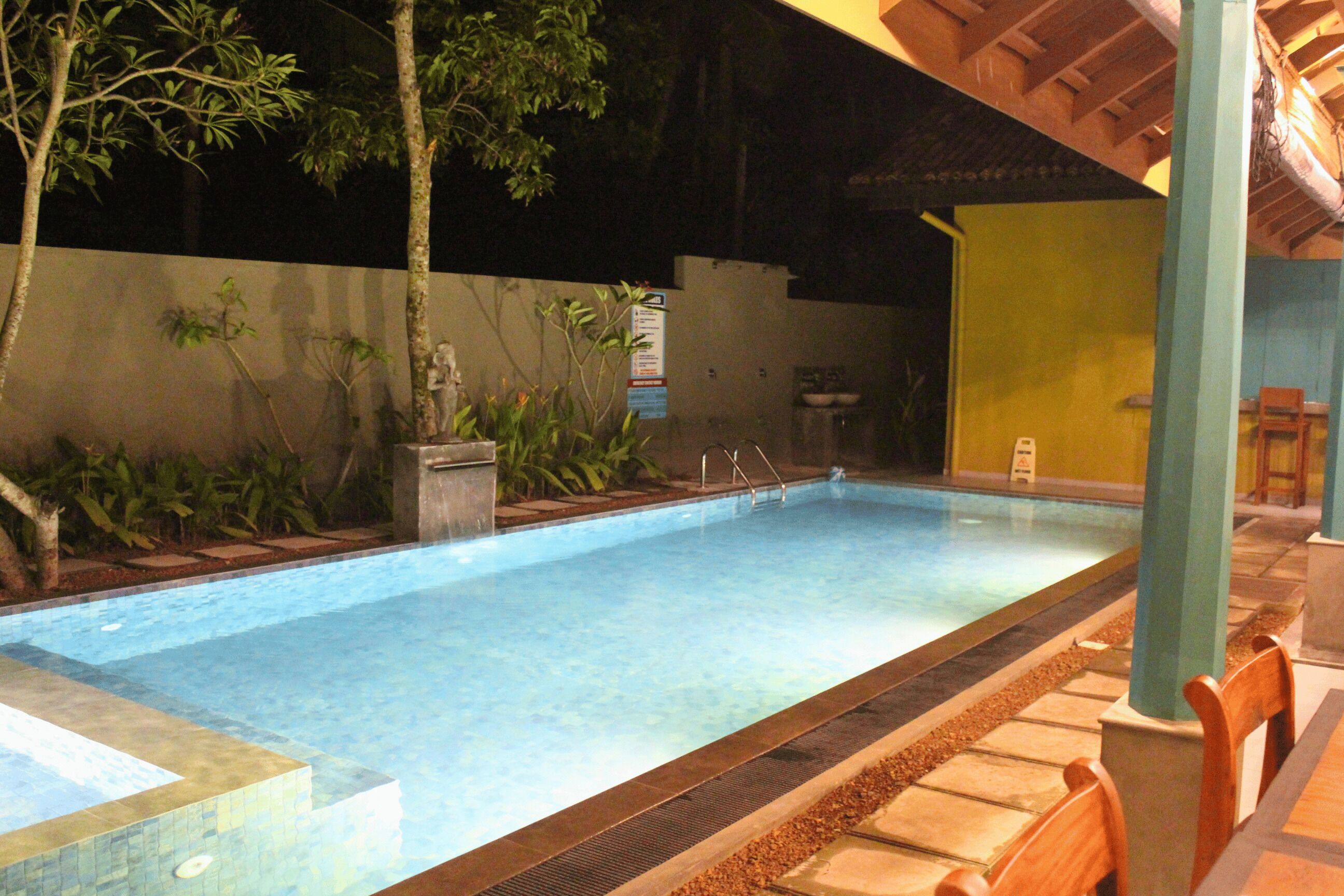 A swimming pool with a tiled edge, surrounded by plants and a covered seating area with chairs.