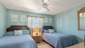 A bedroom with two beds, a ceiling fan, a nightstand with a lamp, and artwork on the walls. - Beachside Breeze 1BR Condo with Pool & Great View! (Wildwood Crest)