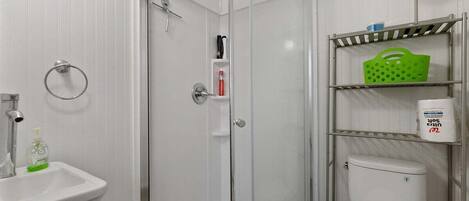 Un baño con ducha acristalada, lavabo, inodoro y estantes con artículos de aseo.