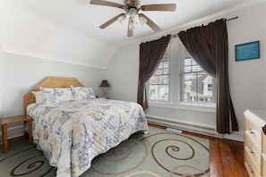 A bedroom with a bed, a nightstand, a ceiling fan, a window with curtains, and a sign on the wall.