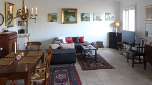 Een woonkamer met een bank, eettafel en stoelen. Er hangen schilderijen aan de muur en er is een kroonluchter.