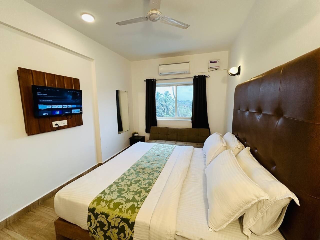 Una camera d'albergo con un letto, una ventola a soffitto, un condizionatore d'aria, una TV fissata al muro e una finestra con tende.