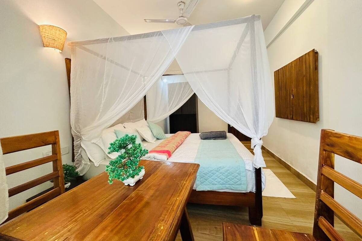 A bedroom with a bed under a canopy, a wooden table with a plant, and a wall-mounted light fixture.