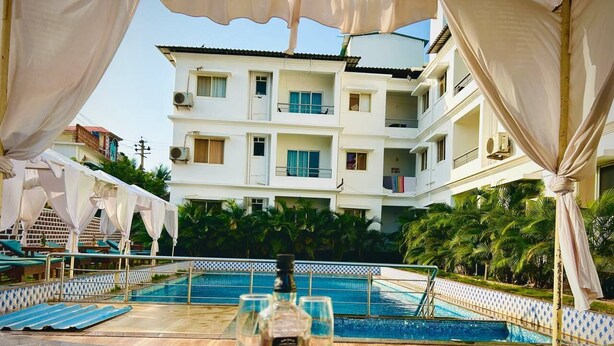 A poolside table with a bottle and glasses, a pool, and a building with balconies in the background. - Dacha Arambol (Arambol)