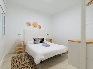 Quarto com cama, mesas de cabeceira, luminária na parede, cesta decorativa de fibra e tapete estampado.