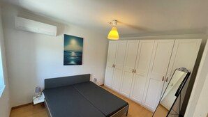 Een slaapkamer met een bed, garderobe, nachtkastje, lamp, airconditioning en een schilderij van een zeegezicht.
