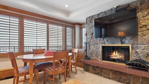 A cozy living room with a stone fireplace, a dining table with chairs, and large windows with blinds. - Lodge Tower 686 2BR at the base of Vail Mountain (Vail)