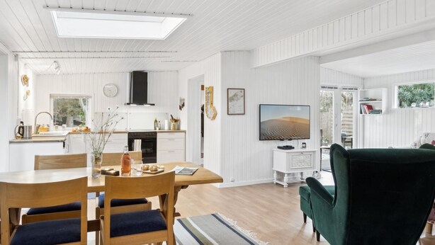 Eine moderne Küche mit weißen Schränken, einem Essbereich mit Holzsesseln, einem grünen Sofa und einem an der Wand montierten Flachbildfernseher.