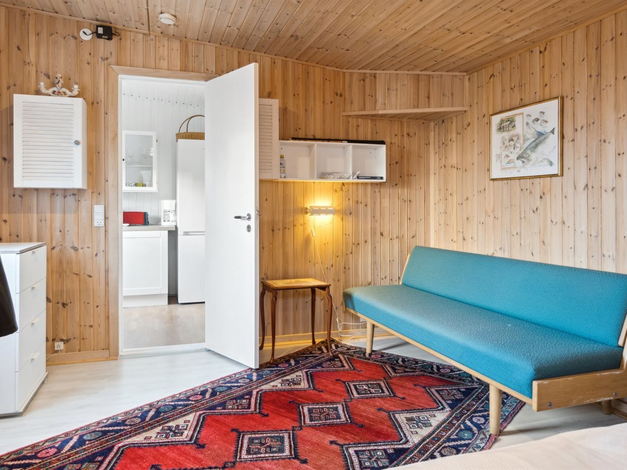 Une pièce aux murs en bois, avec un canapé bleu, un tapis à motifs, une petite table et un coin cuisine visible à travers une porte ouverte.