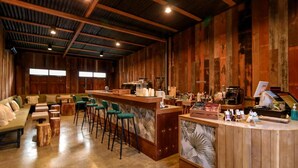 A rustic bar area with wooden walls, a bar counter, stools, and a seating area with a sofa and chairs. - Skugga Farm Cabin (Mae On)