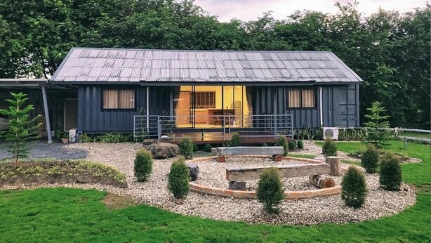 A rustic cabin with a large glass door entrance, surrounded by a well-maintained garden with a circular stone feature and small trees. - Skugga Farm Cabin (Mae On)