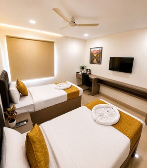Quarto de hotel com duas camas, uma escrivaninha, uma cadeira, uma televisão e um ventilador de teto.