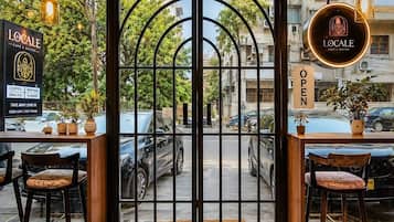 A cafe entrance with a geometric patterned floor, two wooden tables with chairs, and a view of parked cars and buildings outside.