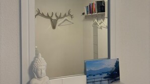 A room with a white wall, a white shelf with a Buddha statue, a framed picture, and a mirror reflecting a ceiling light and a bookshelf.