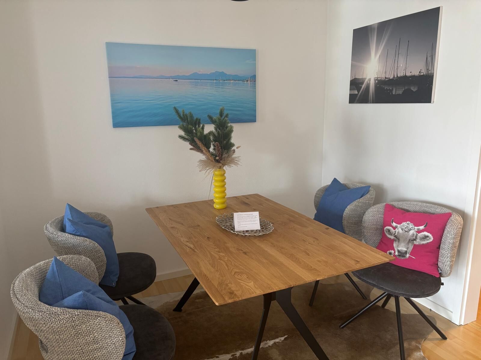 A dining area with a wooden table, two chairs, a vase with dried plants, and two paintings on the wall.