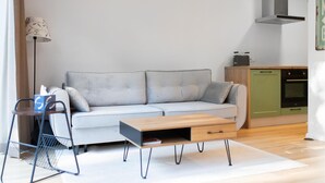 Ein modernes Wohnzimmer mit grauem Sofa, einem Holztisch, einem Metallstuhl und einer Küchenzeile mit einem grünen Schrank und einem Ofen.