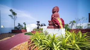 Una estatua de Buda en un jardín con paredes rojas y blancas, plantas verdes y cielo despejado.