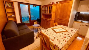 A compact living space with a sofa, a dining table with a framed picture, a kitchen area, and a view through a large window. - Cozy apartment with terrace, parking and mountain view in La Clusaz (La Clusaz)