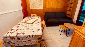 A small, cozy room with a dining table covered in a patterned tablecloth, a black sofa, a wooden bookshelf, and a window with a view. - Cozy apartment with terrace, parking and mountain view in La Clusaz (La Clusaz)