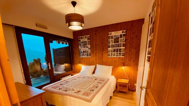 A bedroom with a wooden floor, a bed with a patterned bedspread, a bedside table, a nightstand, a wall-mounted lamp, and a poster on the wall. - Cozy apartment with terrace, parking and mountain view in La Clusaz (La Clusaz)
