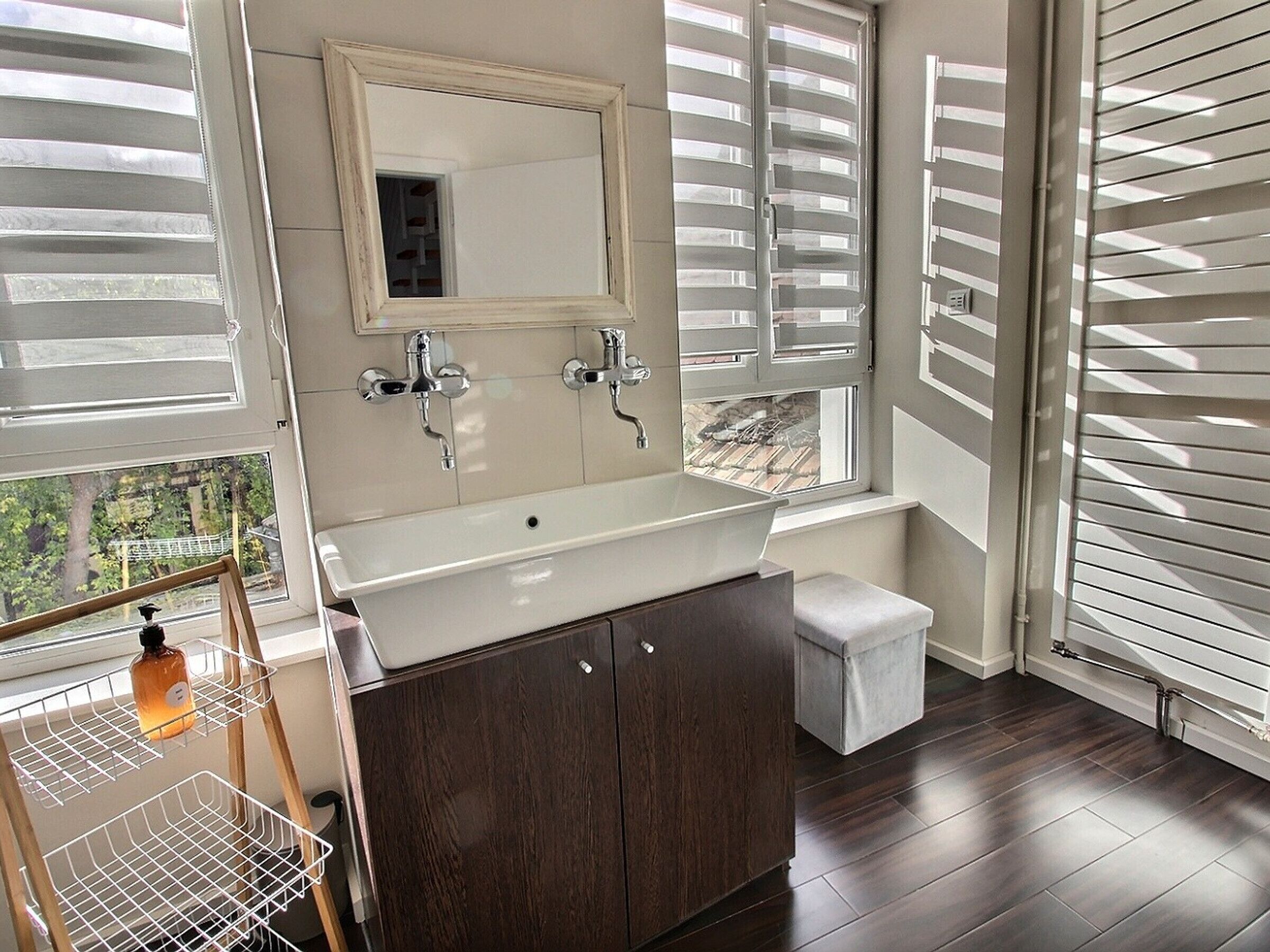 A bathroom with a large window, a white sink, dark wood cabinets, and a small stool.