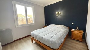 Un dormitorio con cama, mesita de noche, lámpara de pared y ventana con vista a una casa.