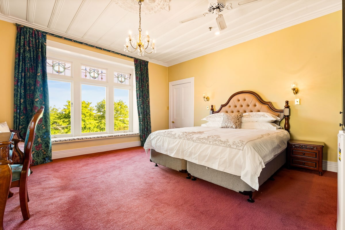 A bedroom with a large bed, a chandelier, a window with curtains, and a view of greenery outside.