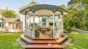 Un mirador con techo, mobiliario exterior y vista a una casa al fondo.
