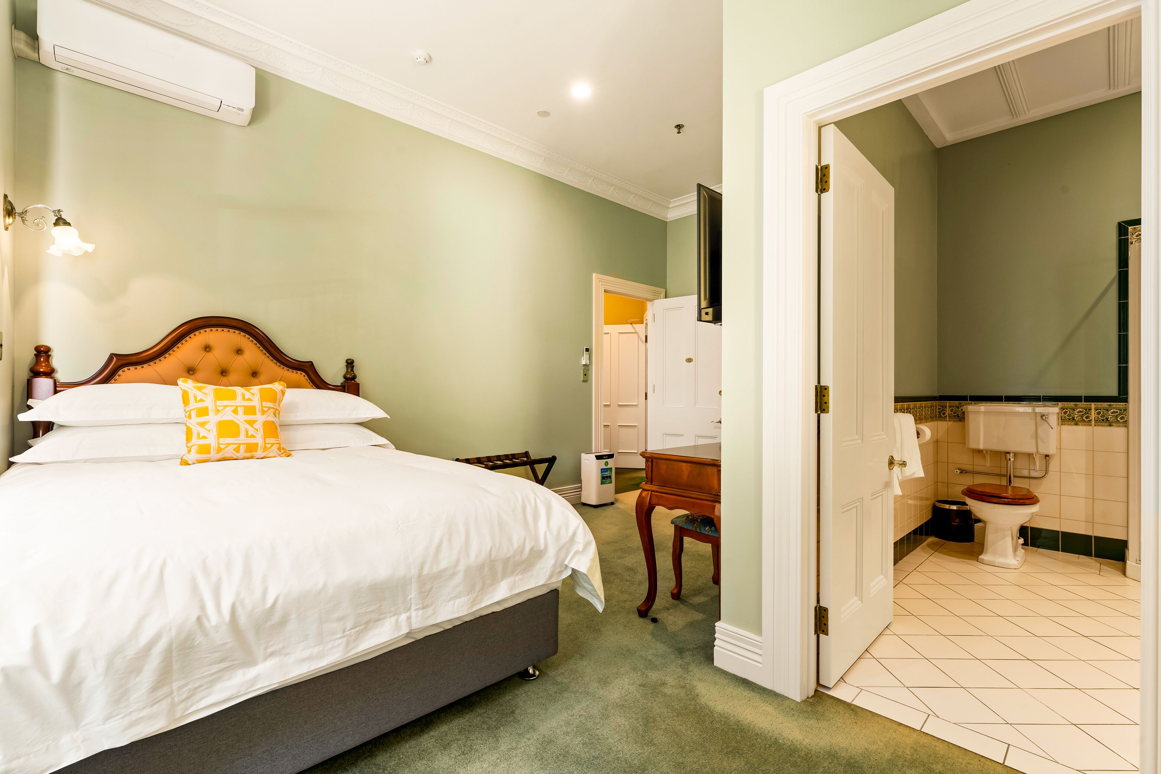 Un dormitorio con cama, mesita de noche, silla, escritorio y baño con inodoro y lavamanos.