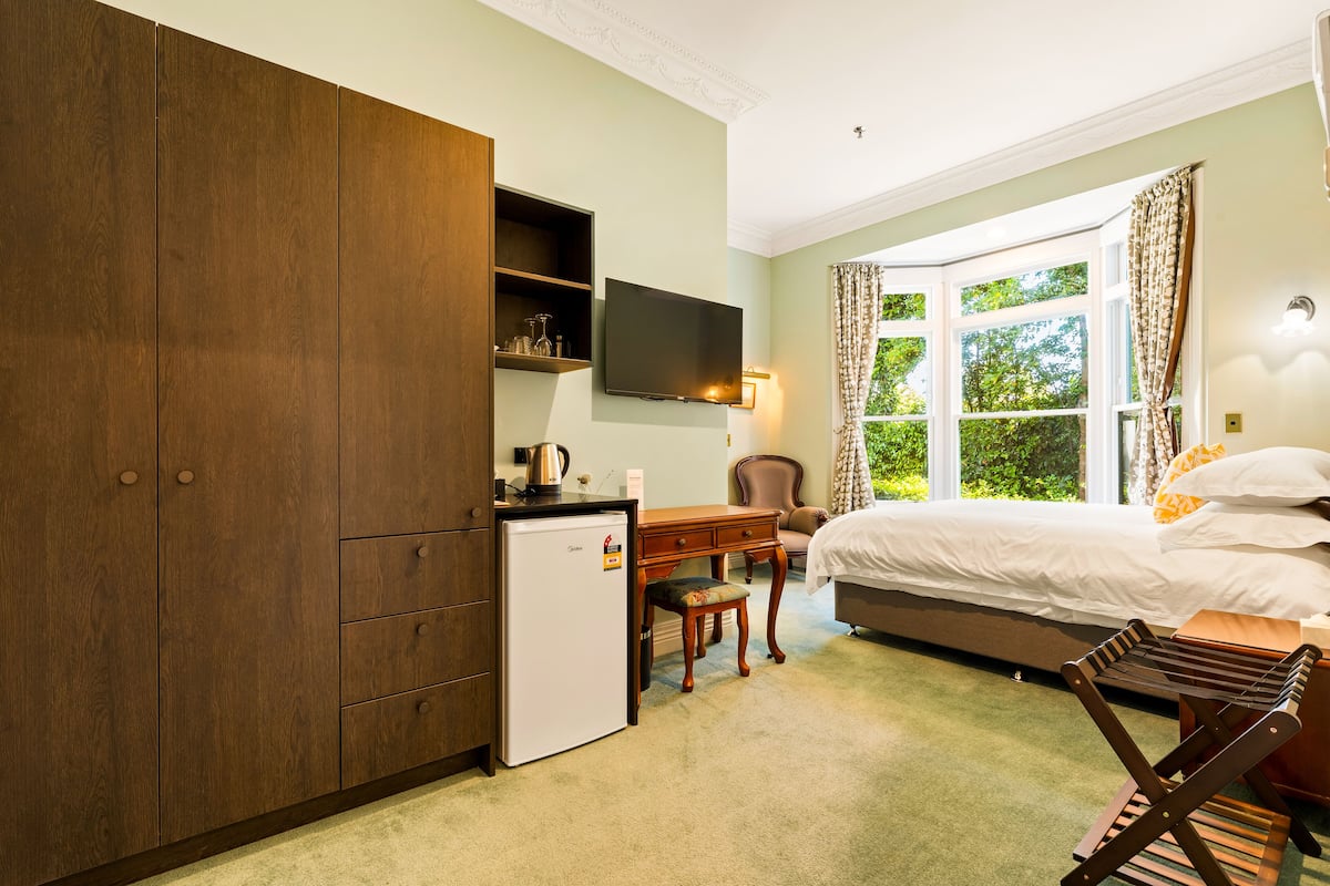 A bedroom with a bed, a desk, a small refrigerator, a television, and a window with curtains.