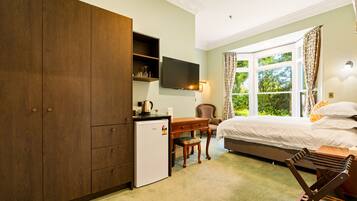 A bedroom with a bed, a desk, a small refrigerator, a television, and a window with curtains.