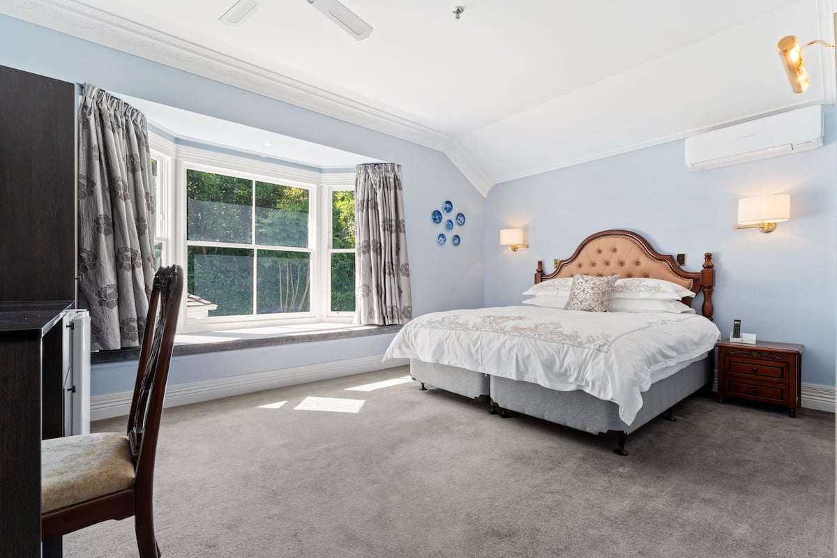 A bedroom with a large bed, a window with curtains, a bedside table, and a wall-mounted lamp.